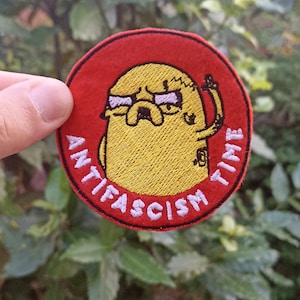 Può includere: Una toppa rotonda rossa ricamata con un personaggio dei cartoni animati giallo che indossa gli occhiali e alza il pugno. Il testo "Anti-fascism Time" è scritto in bianco attorno al personaggio.