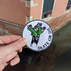 Può includere: Toppa rotonda bianca con pattini a rotelle verdi e il testo "Skate or Die" in nero.