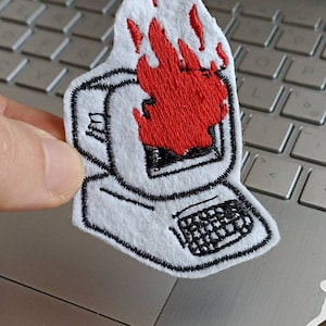 Può includere: Una toppa ricamata che raffigura un monitor di computer con fiamme che escono dallo schermo. La toppa è su un laptop argentato.