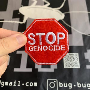 Può includere: Una toppa rossa, ottagonale, con testo ricamato bianco che recita "STOP GENOCIDE". La toppa è tenuta su uno sfondo nero con una grafica di insetti bianca e testo. La toppa è un oggetto di protesta.
