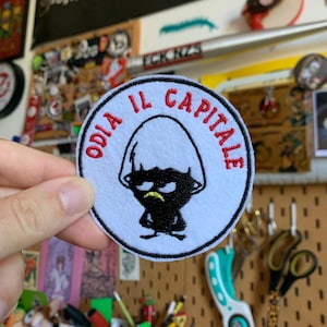 Può includere: Una toppa rotonda bianca con le parole "ODIA IL CAPITALE" ricamate in rosso. La toppa presenta un personaggio dei cartoni animati nero con un'espressione scontrosa e una smorfia gialla.