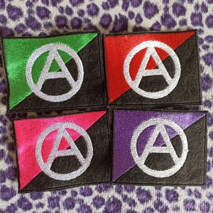 Può includere: Quattro toppe ricamate con il simbolo dell'anarchia in un cerchio. Le toppe sono verde e nero, rosso e nero, rosa e nero e viola e nero.