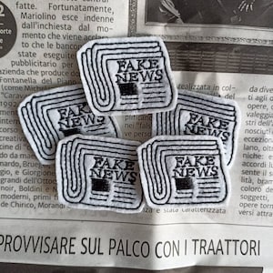 Può includere: Cinque toppe ricamate con le parole "Fake News" in testo nero su sfondo bianco. Le toppe hanno la forma di un giornale piegato.