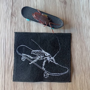 Può includere: Una toppa in feltro nero con ricamo bianco di uno scarafaggio che fa skateboard. Lo scarafaggio ha lunghe antenne ed è su uno skateboard con ruote.
