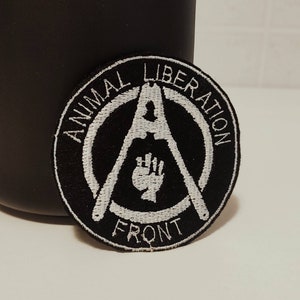 Può includere: Toppa ricamata in bianco e nero con il testo "Animal Liberation Front" e un simbolo anarchico con un pugno chiuso al suo interno.