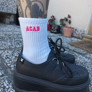 Può includere: Calzini bianchi con la parola "ACAB" ricamata in rosa sulla caviglia. I calzini sono indossati con scarpe platform nere.