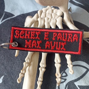 Può includere: Portachiavi in tessuto nero e rosso con il testo "SEHER E PAURA  MAI AVUI" in rosso.