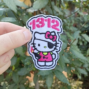 Può includere: Una toppa ricamata bianca con un personaggio di Hello Kitty dei cartoni animati che indossa una benda da pirata e tiene un fiammifero acceso. La toppa ha il testo "1912" sopra il personaggio.