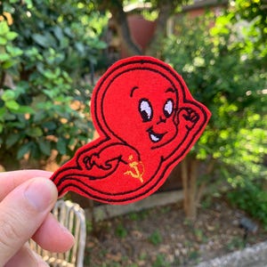 Può includere: Una toppa in feltro rosso a forma di fantasma dei cartoni animati con contorni ricamati neri. Il fantasma presenta un volto sorridente e un piccolo simbolo giallo ricamato di martello e falce sul corpo, con una coda fluida verso sinistra.