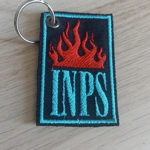 Può includere: Portachiavi nero con un designo a fiamma rossa e la parola "INPS" in lettere blu turchese.