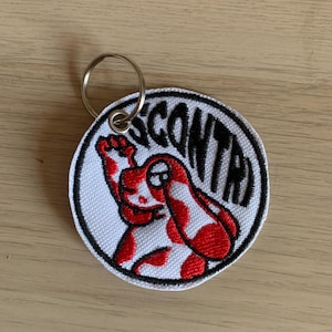 Può includere: Un portachiavi rotondo bianco con bordo nero e la scritta "SCONTA" in nero. Il portachiavi presenta un design di personaggio dei cartoni animati rosso e bianco. È attaccato un anello di metallo argentato.