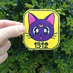Può includere: Un cerotto di feltro giallo con un viso di gatto viola con orecchie rosa e una mezzaluna sulla fronte. Il gatto ha grandi occhi bianchi e una lingua rosa che sporge. Il numero 1312 è stampato in nero sotto il viso del gatto.