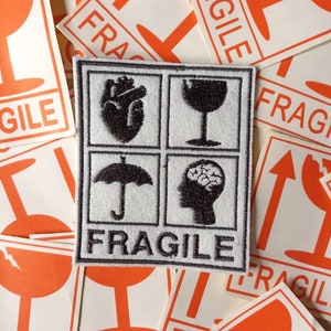 Può includere: Una toppa ricamata in bianco e nero con quattro simboli: un cuore, un bicchiere di vino, un ombrello e un cervello. La toppa ha la scritta "FRAGILE" in nero sotto i simboli.