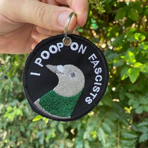 Può includere: Una toppa ricamata in bianco e nero con un uccello verde e il testo "I POOP ON FASCISTS". La toppa è attaccata a un portachiavi argentato.