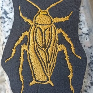 Op de afbeelding: Een gouden geborduurde kakkerlak patch op een zwarte vilt achtergrond.