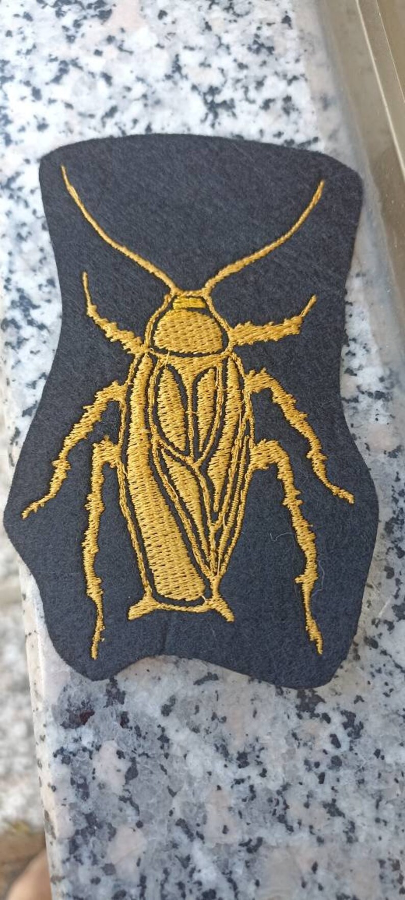 Op de afbeelding: Een gouden geborduurde kakkerlak patch op een zwarte vilt achtergrond.