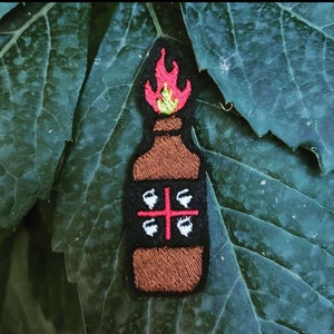 Può includere: Toppa ricamata raffigurante una bottiglia marrone con una fiamma rossa e verde sulla parte superiore. La bottiglia ha un'etichetta nera con quattro disegni di tazze bianche e una croce rossa. La toppa è posta su uno sfondo di foglie verdi.