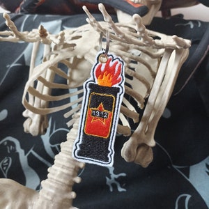 Può includere: Un portachiavi ricamato nero e rosso a forma di cocktail Molotov con fiamme e il numero "1312" sul lato.