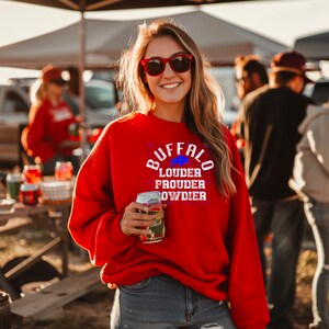 Puede incluir: Una sudadera roja con el texto "BUFFALO LOUDER PROUDER OWDIER" en blanco con una silueta de b&uacute;falo azul. La sudadera la lleva puesta una persona que sostiene una lata de cerveza.
