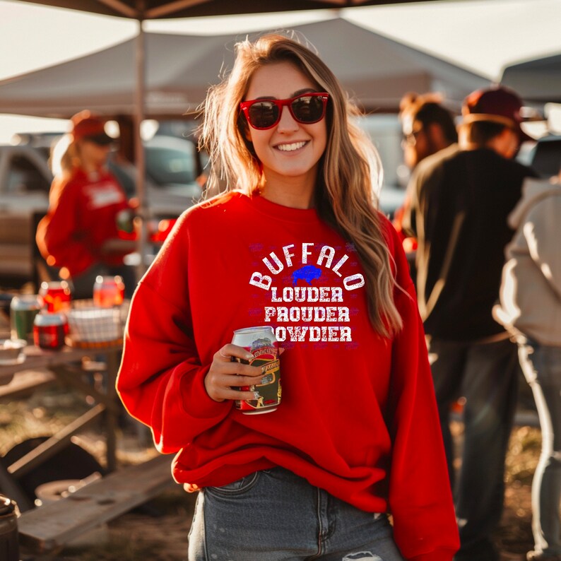 Puede incluir: Una sudadera roja con el texto "BUFFALO LOUDER PROUDER OWDIER" en blanco con una silueta de b&uacute;falo azul. La sudadera la lleva puesta una persona que sostiene una lata de cerveza.