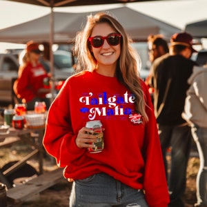 May include: A red sweatshirt with the text "Tailgate Mafia" in white and blue. The sweatshirt also features the "Buffalo Spirit Designs" logo. The person is holding a can of beer and wearing red sunglasses.