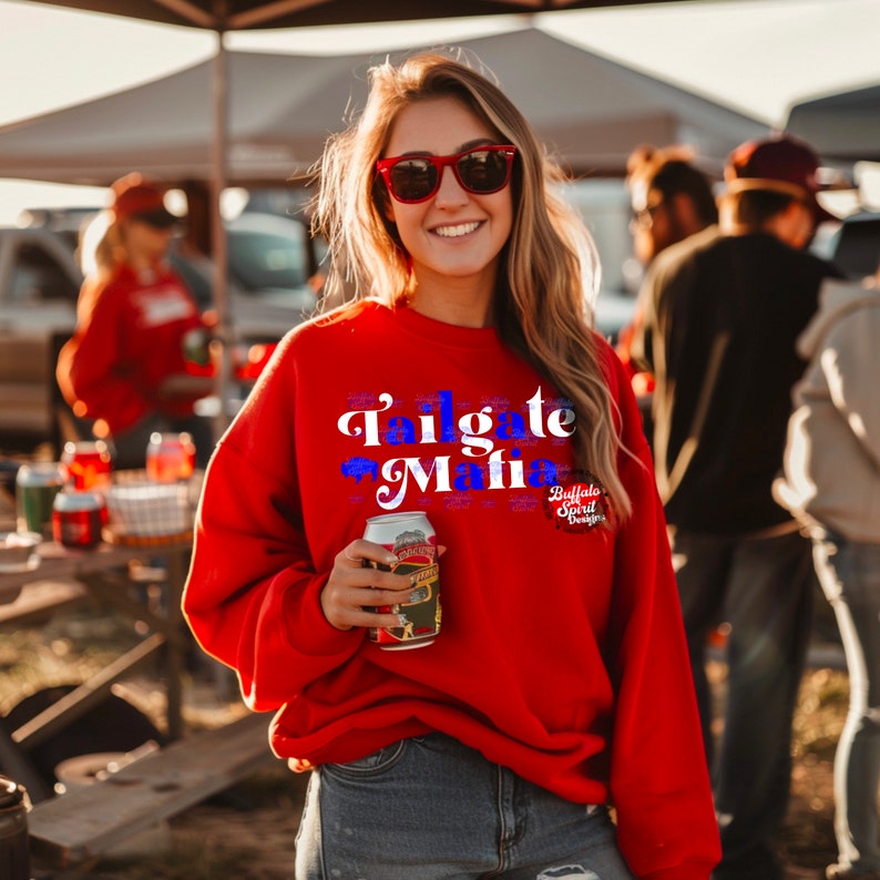 May include: A red sweatshirt with the text "Tailgate Mafia" in white and blue. The sweatshirt also features the "Buffalo Spirit Designs" logo. The person is holding a can of beer and wearing red sunglasses.