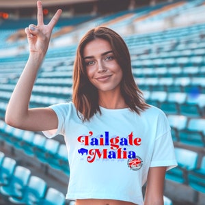 May include: White crop top with the text "Tailgate Mafia" in red and blue, featuring a buffalo graphic. The shirt also has the text "Buffalo Spirit" in a circular design.