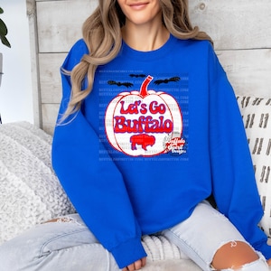 May include: A royal blue sweatshirt with a white pumpkin graphic. The pumpkin has the text "Let's Go Buffalo" in red and blue lettering. The graphic also includes a small buffalo silhouette and the text "Buffalo Spirit Designs".
