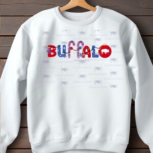 May include: White sweatshirt with the word "BUFFALO" in red, blue, and candy cane-striped letters, decorated with a string of lights and a red ornament. The design includes a buffalo silhouette. The background has a repeating pattern of buffalo heads.