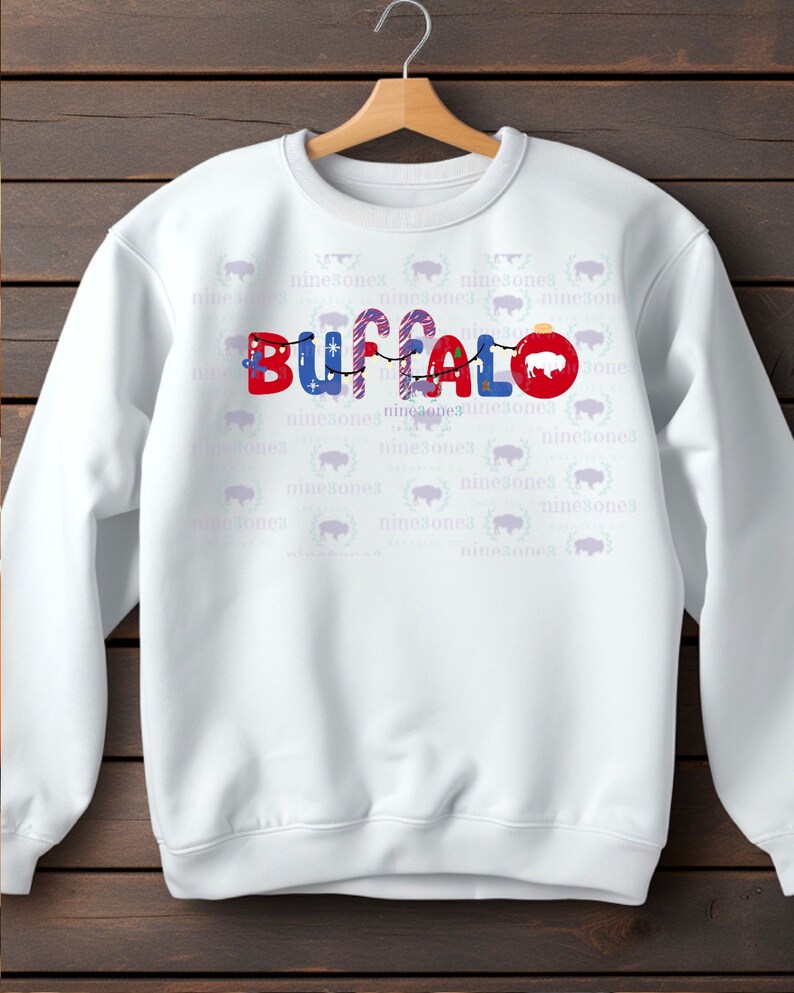 May include: White sweatshirt with the word "BUFFALO" in red, blue, and candy cane-striped letters, decorated with a string of lights and a red ornament. The design includes a buffalo silhouette. The background has a repeating pattern of buffalo heads.