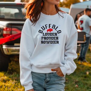 Puede incluir: Sudadera blanca con un dise&ntilde;o gr&aacute;fico desgastado que dice "Buffalo Louder Prouder Rowdier" con una silueta de b&uacute;falo roja.