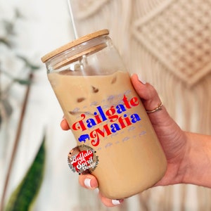 May include: A clear glass tumbler with a wooden lid and straw, filled with a light brown beverage and ice. The words "Tailgate Mafia" are printed in red and blue, with a buffalo graphic. The tumbler is designed by Buffalo Spirit Designs.