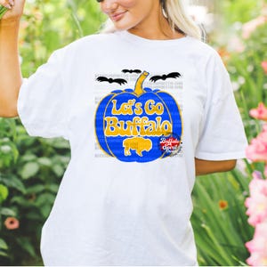 May include: White t-shirt with a blue and gold pumpkin design. The pumpkin has the text "Let's Go Buffalo" on it. There are bats flying around the pumpkin.