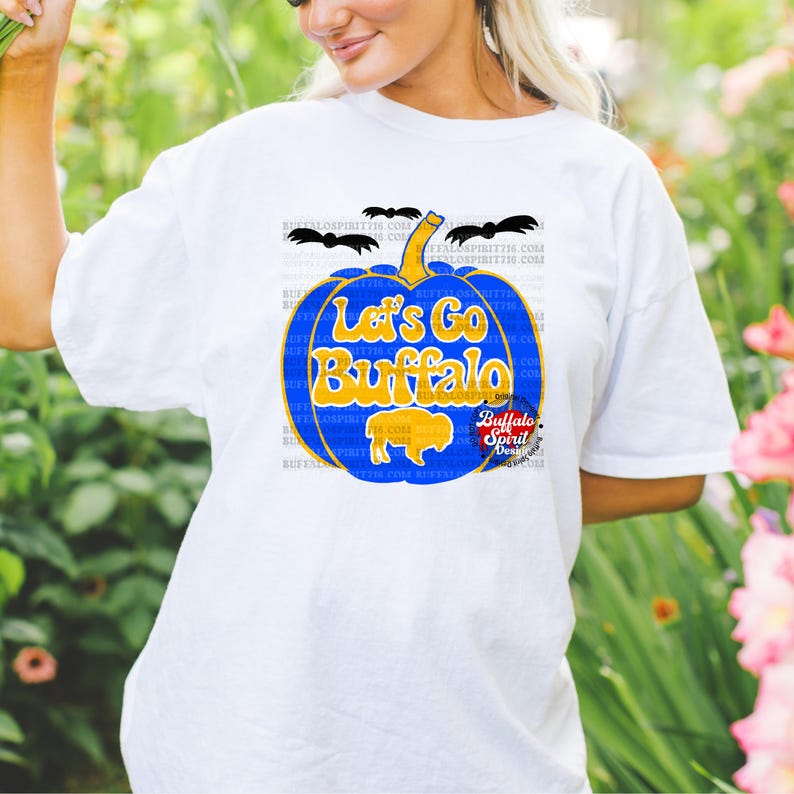 May include: White t-shirt with a blue and gold pumpkin design. The pumpkin has the text "Let's Go Buffalo" on it. There are bats flying around the pumpkin.