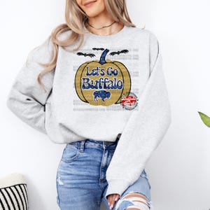 May include: A gray sweatshirt with a glittery gold pumpkin design. The pumpkin has the text "Let's Go Buffalo" on it. The pumpkin is surrounded by bats and the text "Buffalo Spirit" is in a red circle.