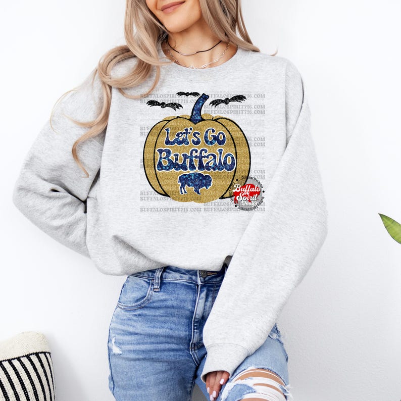 May include: A gray sweatshirt with a glittery gold pumpkin design. The pumpkin has the text "Let's Go Buffalo" on it. The pumpkin is surrounded by bats and the text "Buffalo Spirit" is in a red circle.