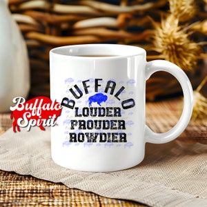Puede incluir: Taza de caf&eacute; de cer&aacute;mica blanca con un dise&ntilde;o en negro y azul. El dise&ntilde;o presenta la palabra "Buffalo" en un dise&ntilde;o circular con las palabras "Louder", "Proud" y "Rowdier" debajo. La taza est&aacute; sobre una superficie de madera marr&oacute;n con un mantel individual de patr&oacute;n blanco y marr&oacute;n.