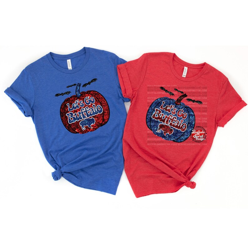 May include: Two t-shirts with a pumpkin design. The pumpkin is decorated with red, white, and blue glitter and says "Let's Go Buffalo". One shirt is blue and the other is red.