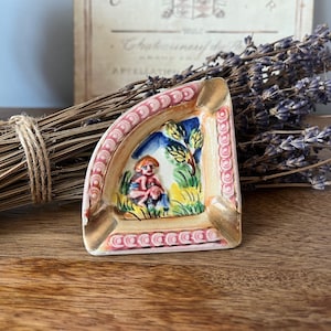 May include: A triangular ceramic ashtray with a pink and gold rim. The ashtray features a hand-painted scene of a woman and a child in a garden setting. The scene is painted in shades of blue, green, yellow, and red.