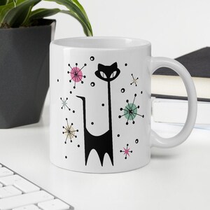 May include: White ceramic mug featuring a black cat design with a long, slender body and a smiling face. The mug is decorated with colorful starbursts in pink, green, and yellow. The mug has a curved handle and is suitable for hot or cold beverages.