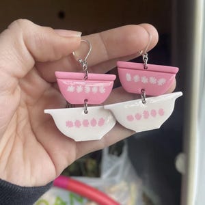 May include: A pair of pink and white ceramic bowl earrings. The earrings are shaped like bowls with a white rim and a pink interior. The bowls have a white floral pattern on the rim.
