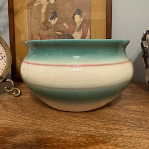 Puede incluir: Un macetero de cerámica vintage con un esmalte verde, blanco y rosa. El macetero tiene una forma redondeada y un borde ancho.