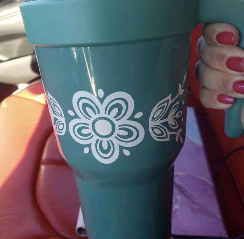 Puede incluir: Una taza de viaje de color turquesa con un dise&ntilde;o floral blanco. La taza tiene un asa y una tapa.