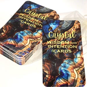 May include: A deck of Crystal Wisdom and Intention Cards with a blue, gold, and brown abstract pattern. The cards are stacked and the top card is visible. The text "Crystal Wisdom and Intention Cards" is printed in gold on the top card.