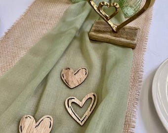 Op de afbeelding: Een gouden hartsculptuur met een kleiner hart erin, op een houten voet. Drie houten hartuitsparingen zijn verspreid over een groene doek. Decoratieve glazen flessen en een vaas op de achtergrond.