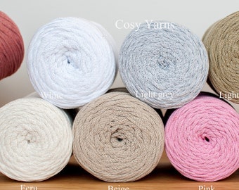 May include: Eight spools of cotton yarn in various colors, including white, light grey, light khaki, ecru, beige, pink, and light lavender. The spools are arranged in a row on a wooden surface. The text "Cosy Yarns" is visible above the spools.