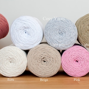May include: Eight spools of cotton yarn in various colours, including white, light grey, light khaki, ecru, beige, pink, and light lavender. The spools are arranged in a row on a wooden surface. The text "Cosy Yarns" is visible above the spools.