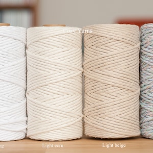 May include: Four spools of cotton rope in different colours: white, light ecru, light beige, and rainbow. The spools are arranged in a row on a wooden surface.