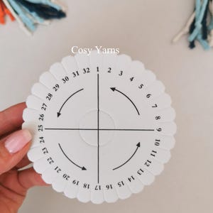 May include: A white, circular yarn tool with scalloped edges, featuring numbers 1-32 and directional arrows. The text "Cosy Yarns" is printed on the tool. The tool is designed for crafting and is held by a person.