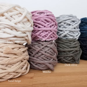 K&ouml;nnte beinhalten: Sortiment von 10 Kn&auml;uelen Baumwollseilgarn in verschiedenen Farben, darunter helles Ecru, Sandbeige, staubiges Rosa, Mokka, Grau, Dunkeloliv, Petrolblau und Dunkelblau. Das Garn ist mit "Cosy Yarns" gekennzeichnet.